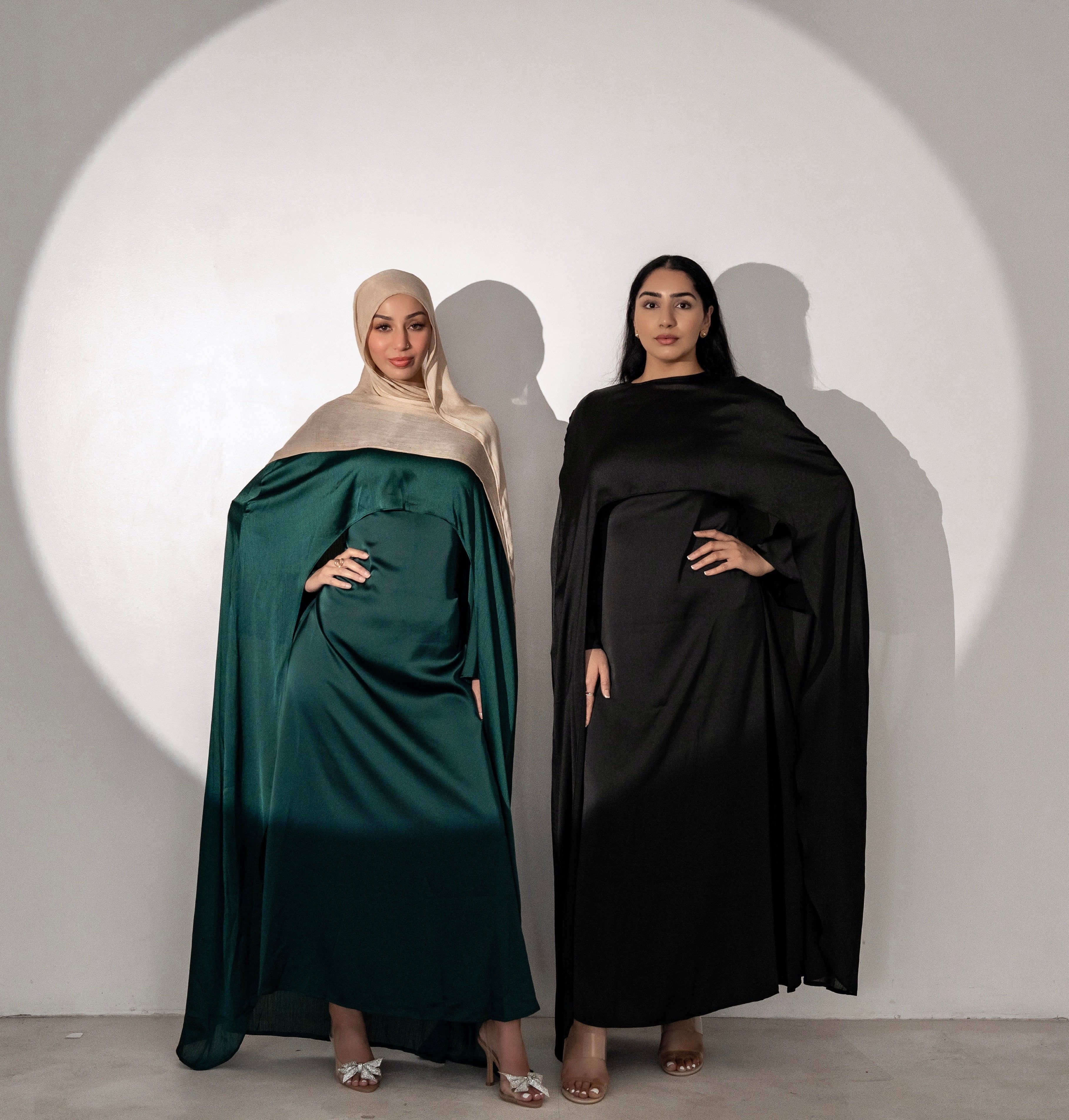 Two women wearing abayas in green and black standing against a white background